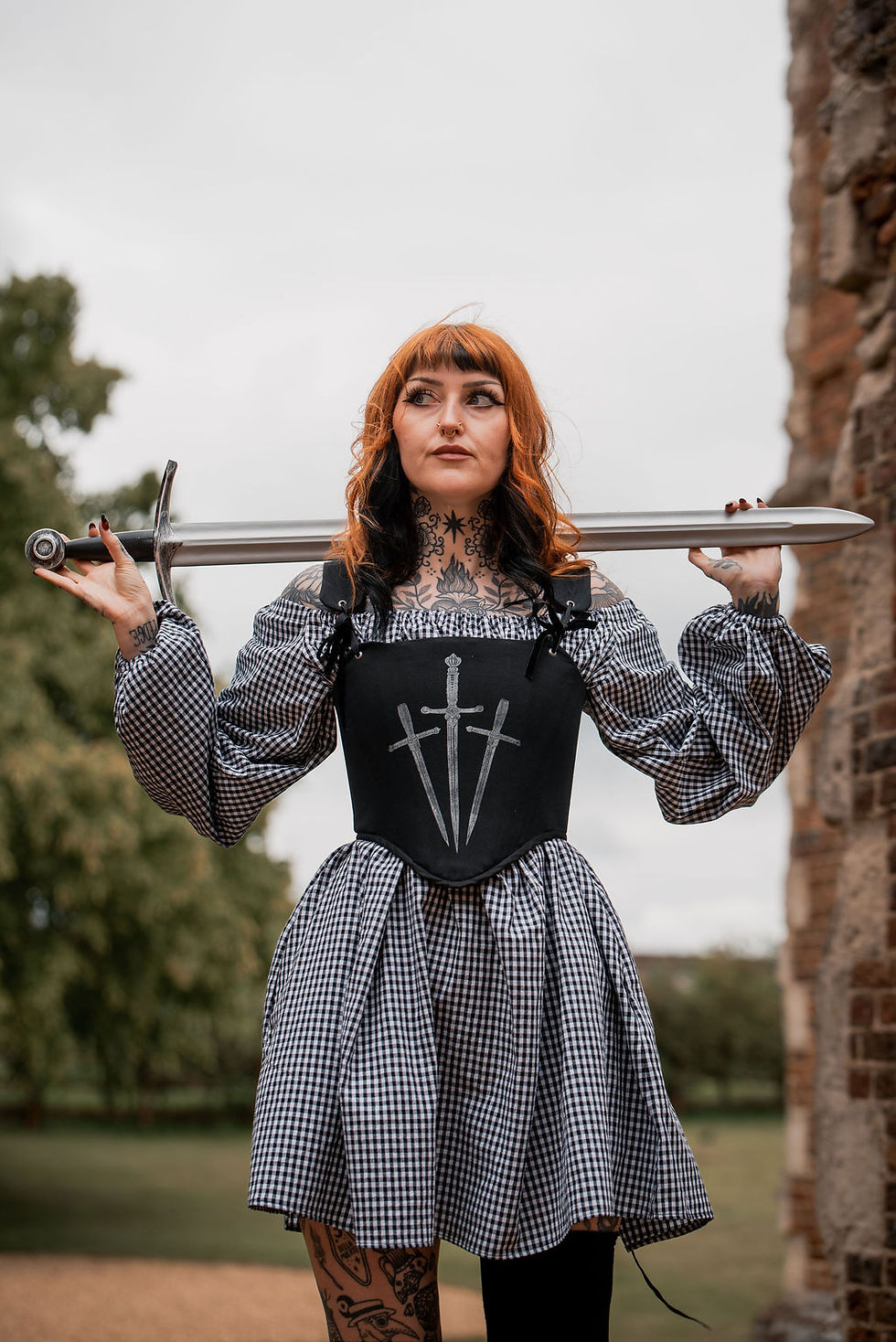 Thumbnail: a tattooed model wearing a gingham dress with black corset holding a sword