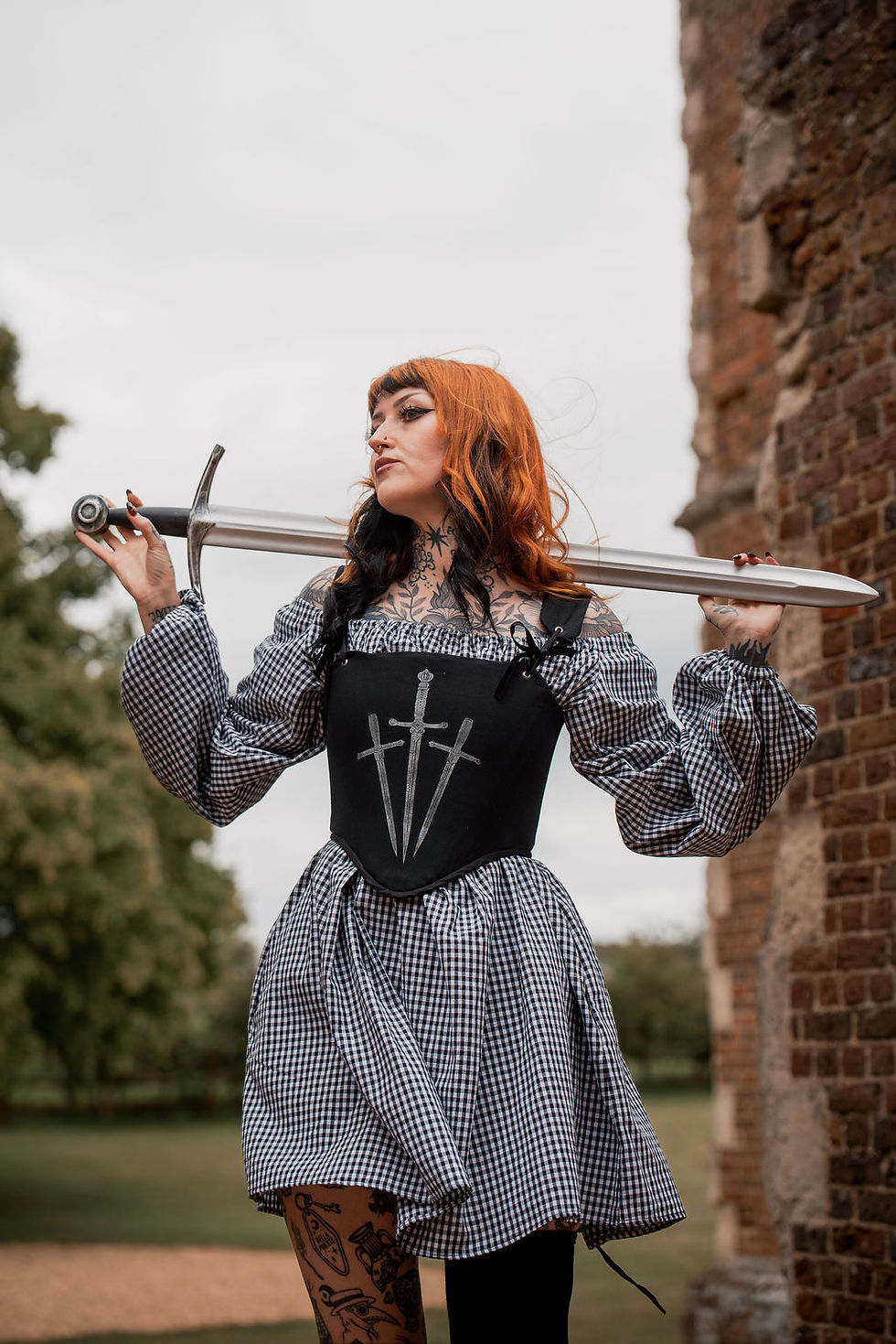 a tattooed model wearing a gingham dress with black corset holding a sword in some ruins