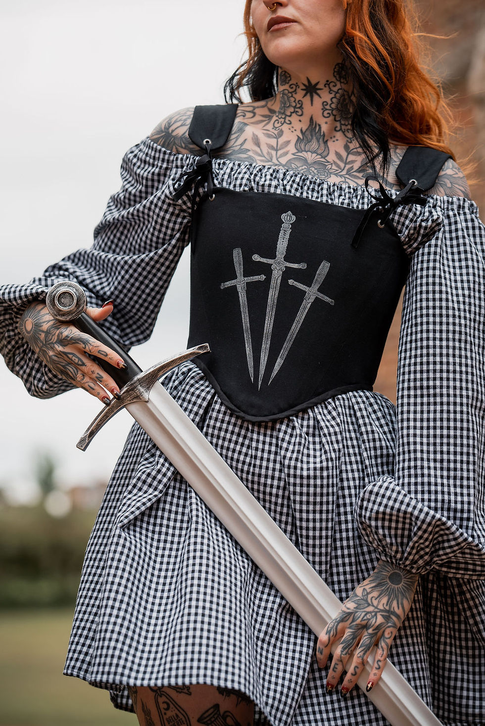 a tattooed model in a gingham dress with black corset with sword print holding a sword