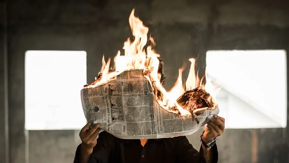 Person holding a burning newspaper in front of their face, flames rising. Background shows a dim room with rectangular windows. Dramatic mood.