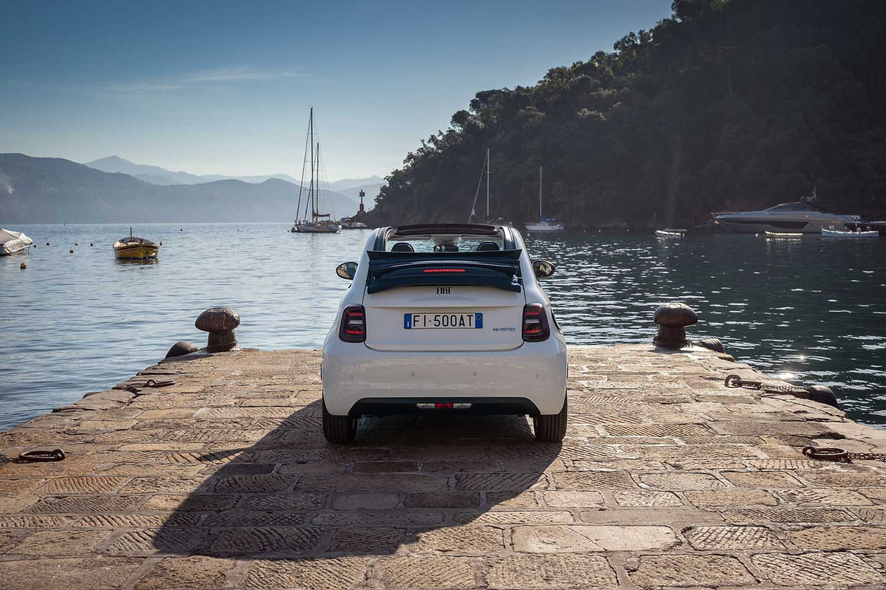 Vista posteriore di una Fiat 500 bianca cabrio parcheggiata su un molo in pietra sul mare. Sullo sfondo, barche e velieri sono ormeggiati in acque tranquille, circondate da montagne e vegetazione, creando un tipico paesaggio costiero italiano.