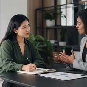 Duas profissionais em uma conversa de desenvolvimento no ambiente de trabalho, sentadas frente a frente em uma mesa de escritório. Uma delas fala e gesticula com naturalidade, enquanto a outra escuta com atenção e faz anotações em um caderno. A cena transmite aprendizado, abertura ao feedback e desenvolvimento profissional, em um ambiente corporativo moderno, com iluminação suave e detalhes em tons neutros e verde.