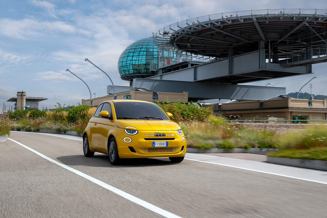 Fiat 500 Hybrid gialla in movimento su una strada urbana moderna di Torino, con architettura contemporanea sullo sfondo.