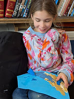 young girl sitting in a floor chair, smiling while looking at her school work on her lap,