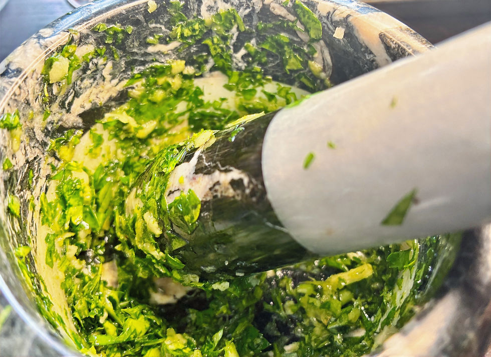 White pestle crushes chopped parsley and garlic in a stone mortar, making a bright pesto-like paste.