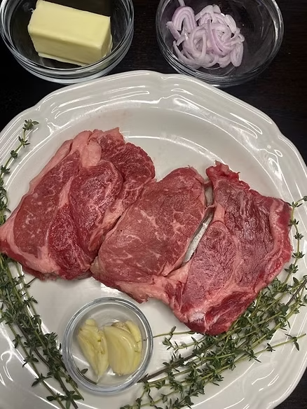 Raw beef steaks on a white plate with thyme and garlic, plus butter and sliced shallots in bowls on a dark table