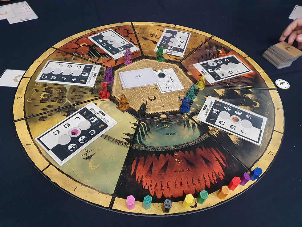 The Veield Fate game board. It is circular and divided into 12 number segments and then a thirteen segment with 'The Pools' and 'The Abyss' in that segment there are a variety of different coloured pieces. There are six cards placed on the board, the first between segments one and two, the second between three and four and so on. The middle of the board is a heptagon and has some facedown cards and one face up card and miniatures can be seen on its edge in various colours. Airecon Veiled Fate - Katenic - March 2023