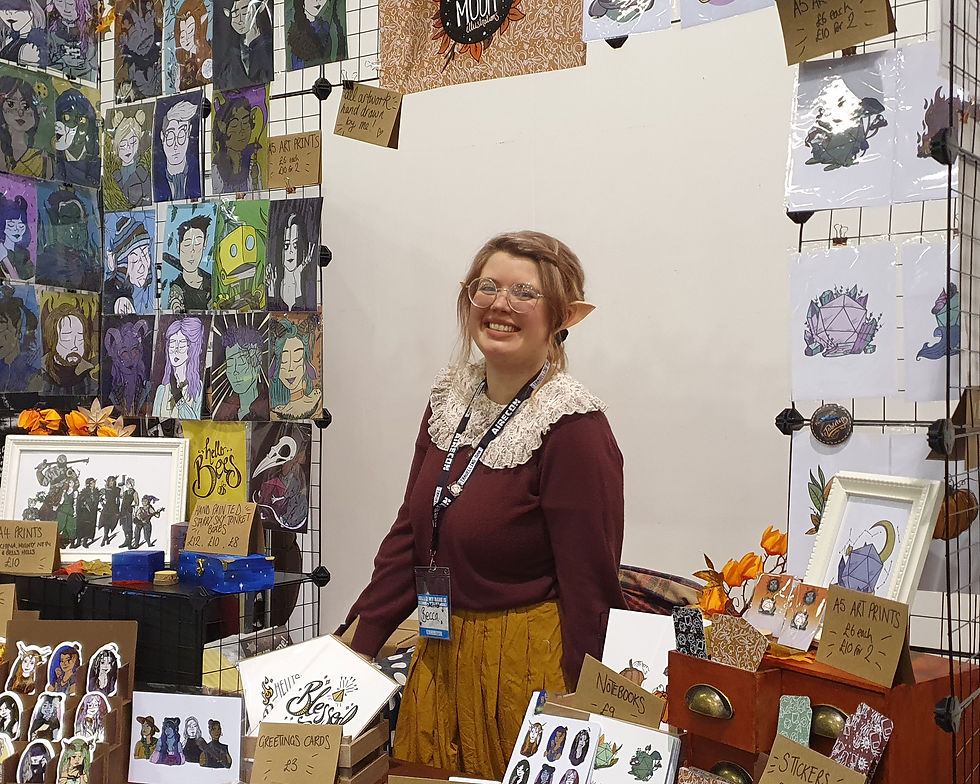 A photograph of Becca from Misty Moon Illustrations; she is wearing a burgundy top with a white laced collar and a light brown skirt and an exhibitor lanyard. She has light coloured hair, round gold-rimmed glasses, elven ears and is smiling as she sands at her stall. The stall has a metal cage with her artwork affixed to it and a table with other items to purchase including notebooks, cards and stickers. Airecon - Misty Moon Illustrations - Katenic - March 2023