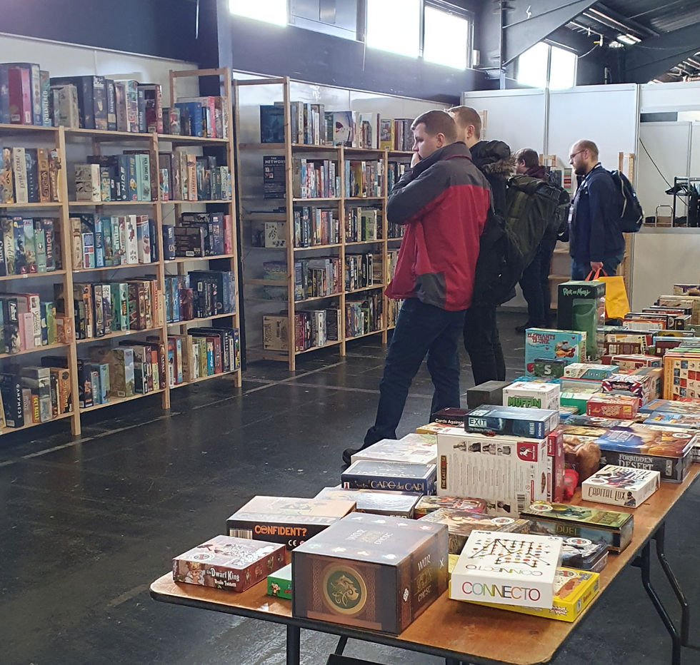 A photograph of the Airecon Games Library where visitors can pick out games to play, there are shelves in the background and a table of games in the foreground. Some visits can be seen browsing. Airecon Games Library - Katenic - March 2023