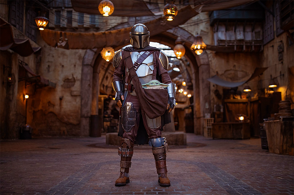 Armored figure stands in a dimly lit, rustic alley, carrying a small creature in a pouch. Warm lights and archways create a mysterious mood.