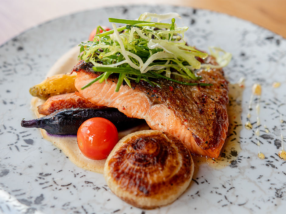 Grilled salmon garnished with greens on a patterned plate, accompanied by cherry tomatoes, roasted onions, carrots, and a creamy sauce.
