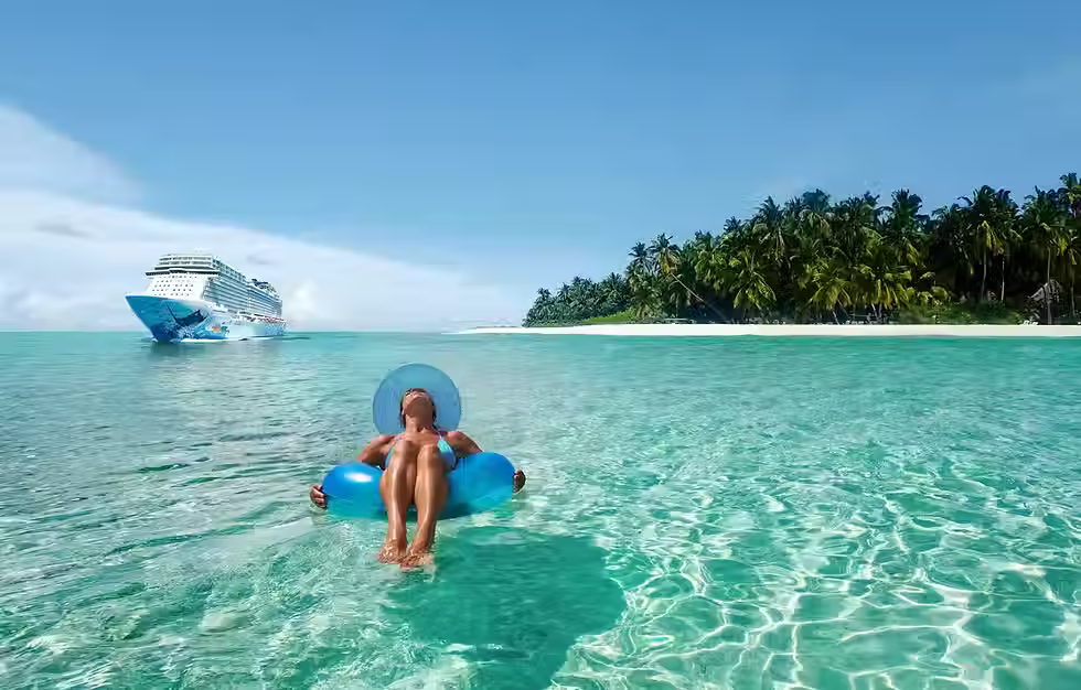 A woman floats on a tube in clear blue water. She is near an island. A Norwegian Cruise Line ship is in the distance.