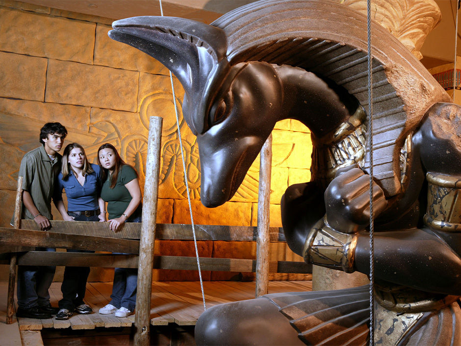 Three people gaze at a large, dark Anubis statue in an Egyptian-themed room. The background features hieroglyphs and warm lighting.