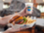 Hands holding a tray with a bacon cheeseburger, fries, and a shake. "Universal Orlando" text visible. Blurred indoor setting.
