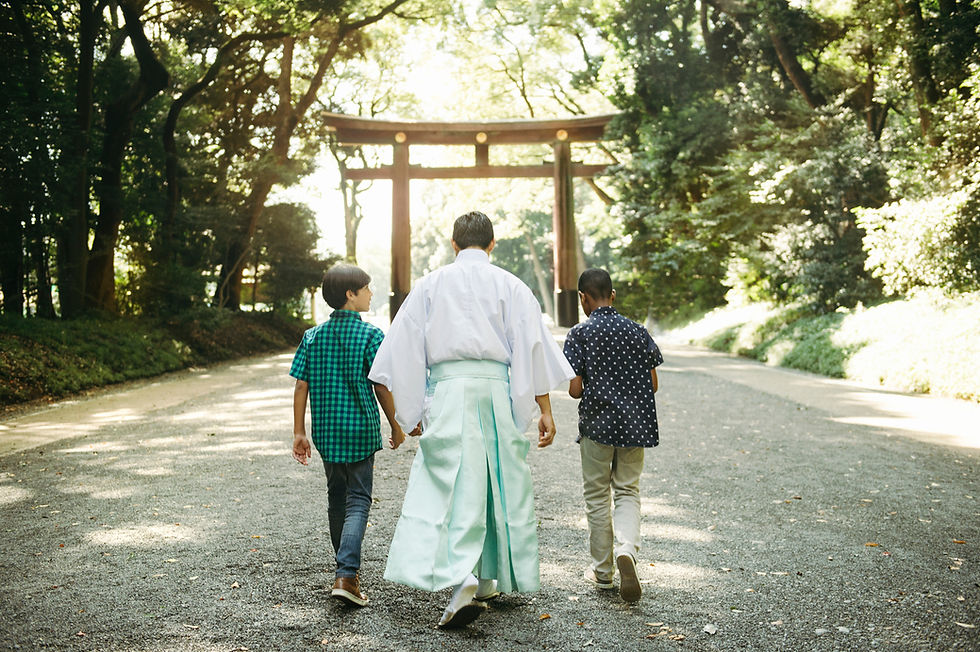 Three people walk on a path towards a wooden torii gate in a forest. The scene is sunlit and serene, with dappled greenery around.