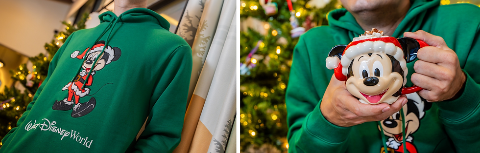 Person in a green Disney World hoodie with Santa Mickey design, holding a Mickey mug with hot cocoa. Christmas tree in background.