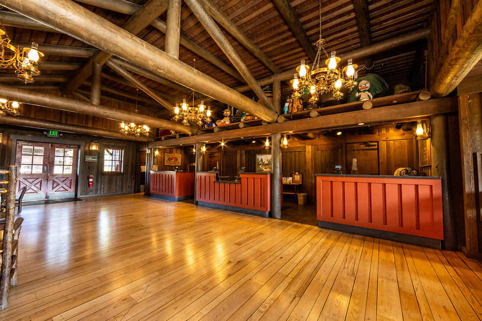 Rustic lodge lobby with wood beams, chandeliers, red front desks, polished floor, and an EXIT sign.