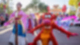 Parade features a person in traditional attire and a red dragon mascot on a sunny day. Colorful costumes and Lunar New Year banner in background.