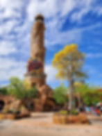 Tower with "Islands of Adventure" sign, surrounded by trees and rocks, under a blue sky. People gather around the base, enjoying the day.