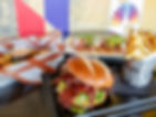 Burger with bacon, fries in a metal cup, and sauce on a table. Colorful wall background. Appealing and inviting food setting.