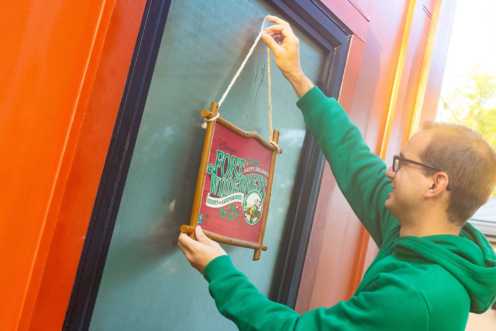 Person in green hoodie hangs a "Fort Wilderness" holiday sign on a glass door. Orange wall in the background, smiling expression.