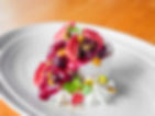 Dessert on white plate with berry sauce, pistachios, and mint leaves. Topped with red fruit pieces. Wooden table background.