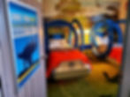 Blue dinosaur-themed bedroom with two circular beds, red blankets. Wall mural of dinosaurs and forest. Sign reads "Dino Food Only." Playful mood.