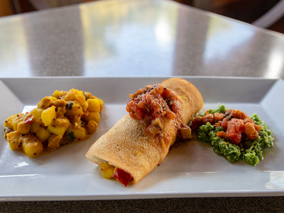 A dosa topped with chutney is plated with spiced potatoes and a green chutney on a rectangular dish. The background is softly blurred.