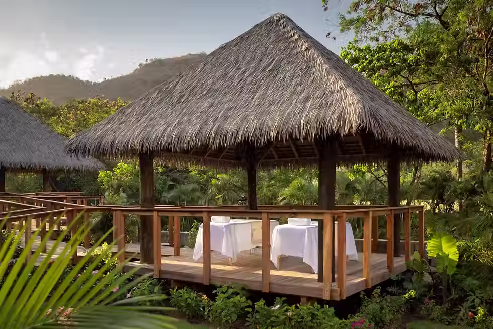 A wooden gazebo with a thatched palm‑leaf roof sits on a raised deck surrounded by lush tropical greenery. Inside are two massage tables covered in crisp white linens, positioned side‑by‑side and facing outward toward the vibrant landscape. Palm trees, flowering plants, and distant green hills create a serene, spa‑like atmosphere perfect for outdoor relaxation.