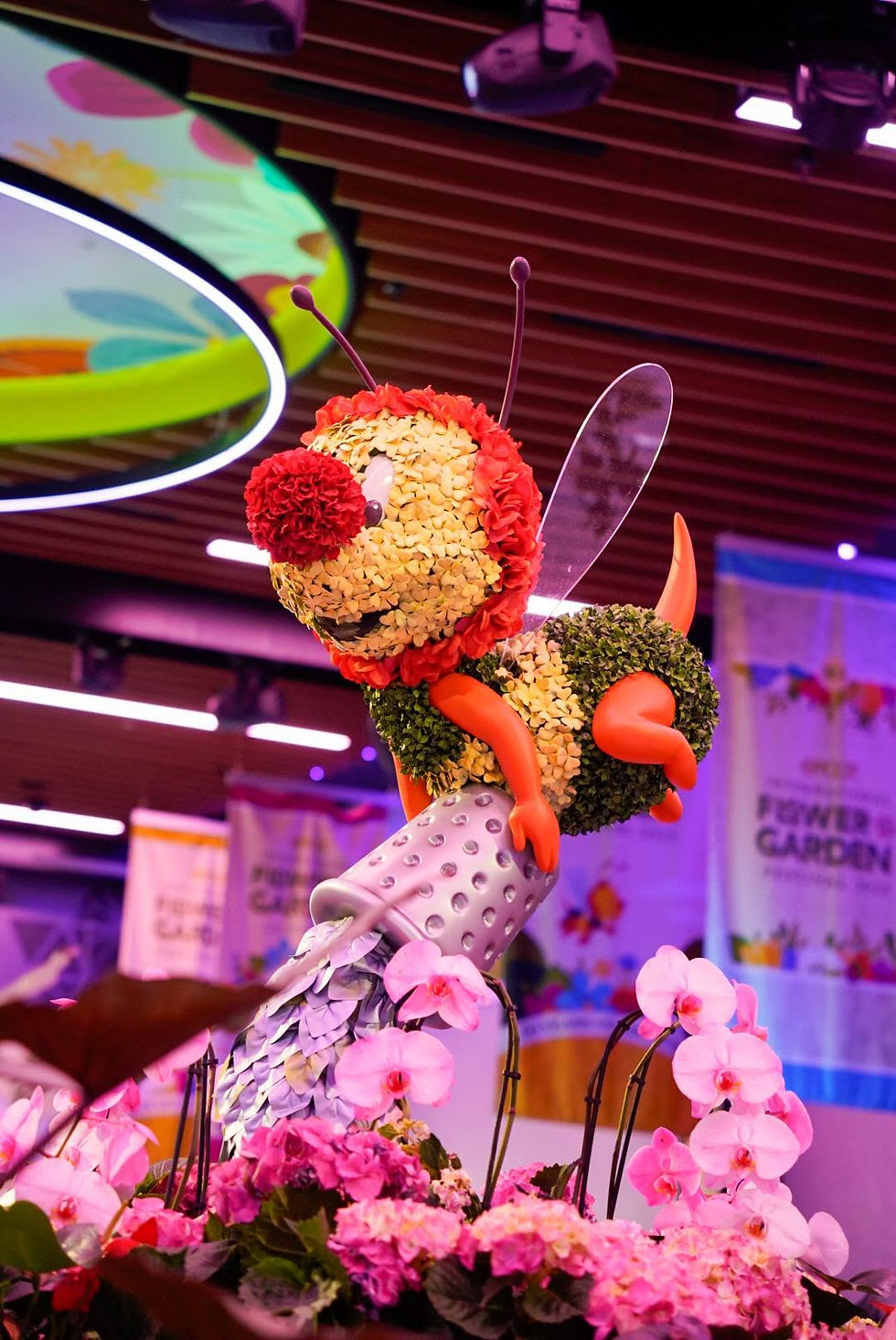 Colorful bee sculpture made of flowers and foliage in an indoor garden display, with blurred Flower Garden banners.