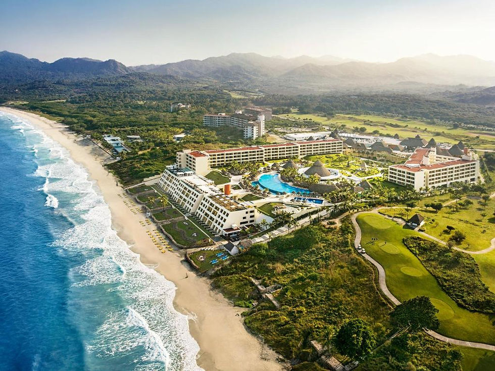 Aerial view of a coastal resort with large white buildings, a blue pool, lush gardens, and a golf course beside the ocean under a sunny sky.