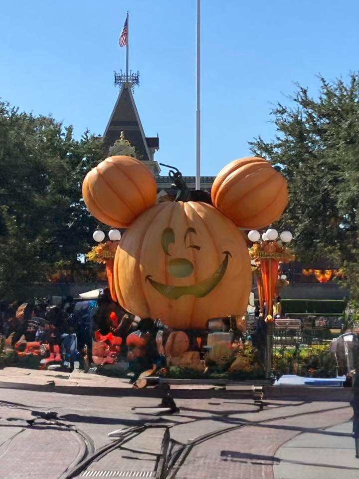Large pumpkin with Mickey Mouse ears and a winking face in a festive park setting. Bright orange and green decorations surround it.