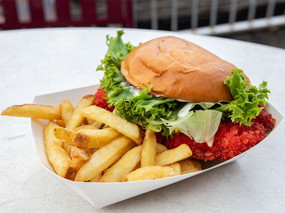 Burger with lettuce, sauce, red crumbed patty in a paper tray with fries. Set on light table outdoors. Bright, appetizing presentation.