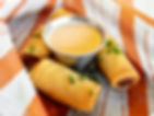 Crispy spring rolls garnished with herbs surrounding a metallic cup of yellow dipping sauce on a patterned orange and white paper.