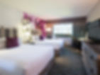 Hotel room with two double beds, white linens, and purple pillows. Floral and geometric wall patterns, large window with daytime view. Cozy mood.