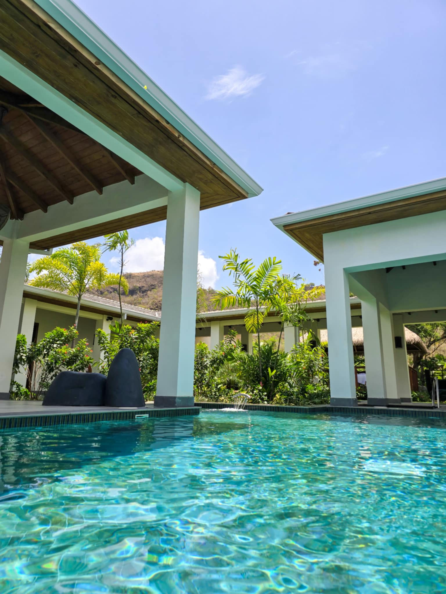 A view from a pool, looking into multiple covered areas. There are green plants and palm trees in the background. The sky is blue.