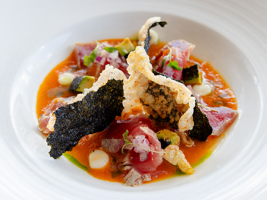 Gourmet dish with tuna, crispy seaweed, and colorful garnishes on a red sauce. Elegant presentation on a white plate.