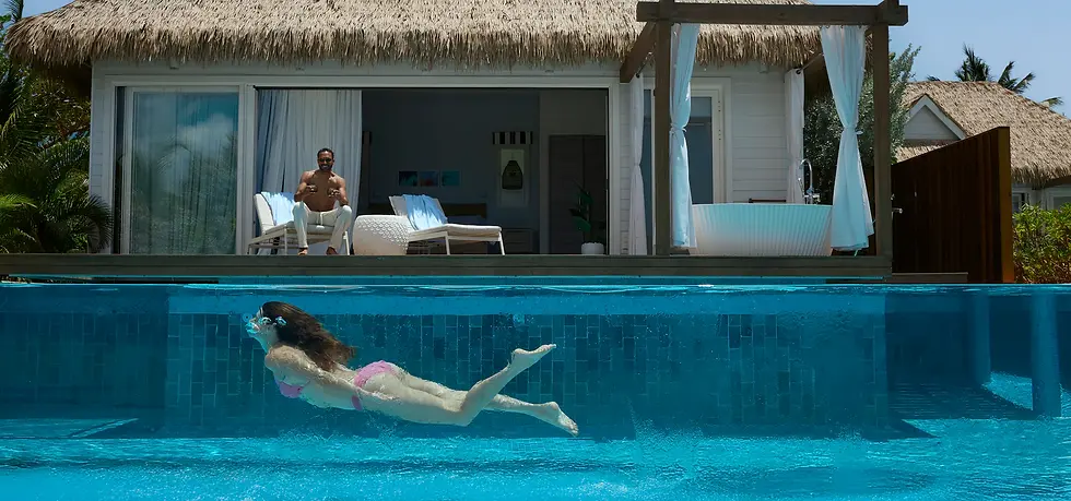 Woman swimming underwater in a clear pool in front of a tropical villa. A man relaxes on a lounger with drinks, sunny, tranquil setting.