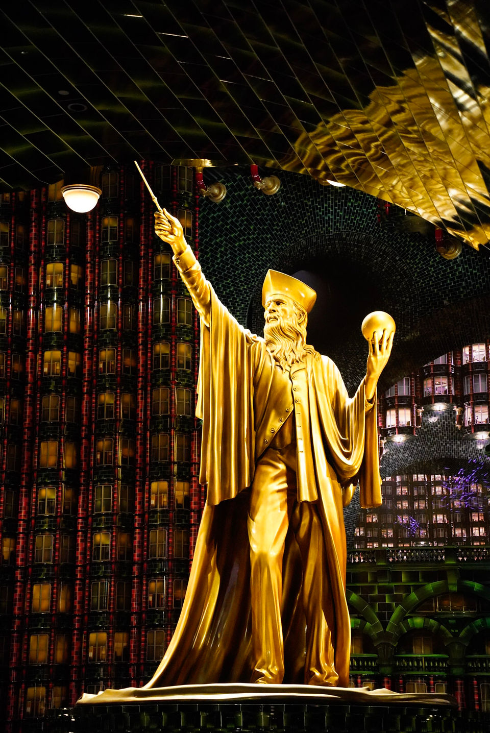 A gold statue of a wizard holding a wand and a ball fills a room. Fake office buildings are behind it.