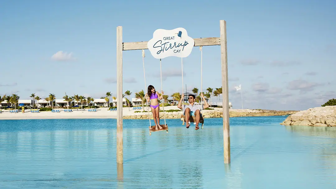 A child and adult are swinging on swings above water. A beach is in the background. A sign reads Great Stirrup Cay.