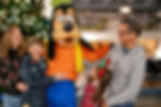 Family posing joyfully with Goofy in festive attire, with Christmas decor in the background. Bright, cheerful atmosphere.