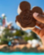 Hand holding a Mickey Mouse-shaped ice cream sandwich against a clear blue sky and blurred green landscape, evoking a joyful, sunny day.
