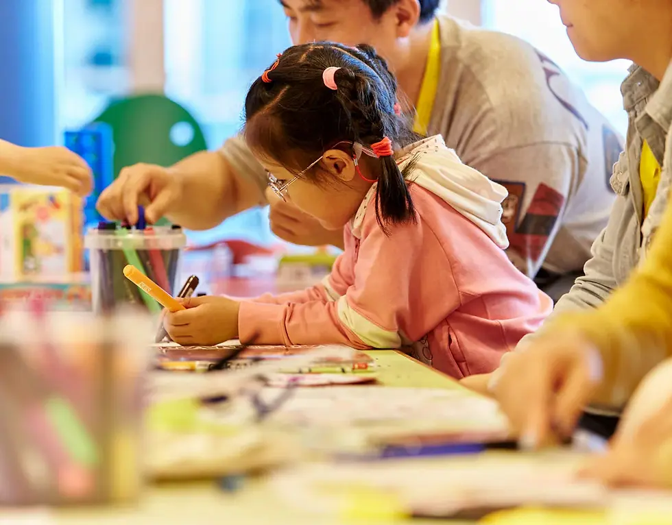 Young girl with glasses colors at a classroom table while adults help nearby, in a warm, focused art activity.