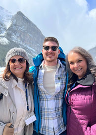 Three people stand in warm clothes in front of a snow-covered mountain. The sky is blue.