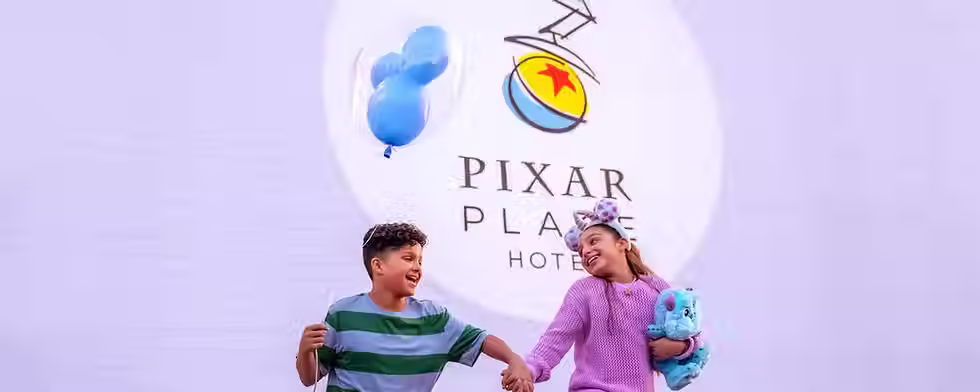Kids are playing outside. One is holding a blue Mickey balloon and the other is holding a Sulley from Monsters Inc. plush. They are standing in front of a sign that says Pixar Place Hotel.