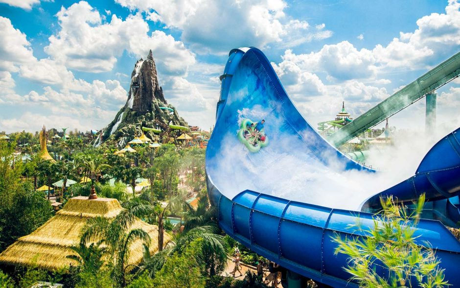 People on a blue water slide at a tropical water park, with a volcano structure in the background. Bright, sunny day with lush greenery.