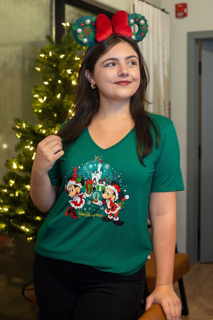 Woman in festive green shirt with Mickey and Minnie, wearing Christmas-themed ears. Smiling by a lit Christmas tree indoors.