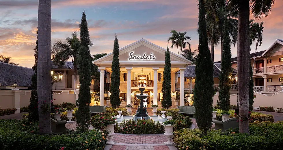 Elegant resort entrance with palm trees and fountain, sunset sky. "Sandals" sign above grand entrance, warm lights create inviting glow.
