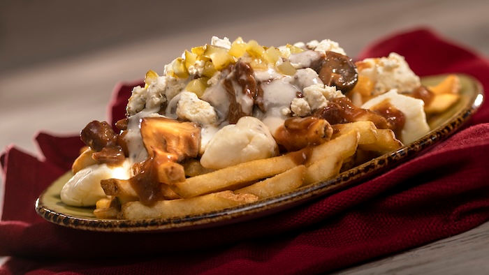 Poutine with fries, cheese curds, gravy, and toppings on a dark plate over a red cloth. The dish looks savory and delicious.
