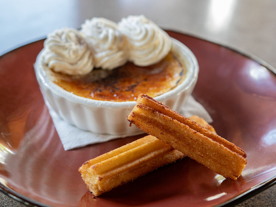 Crème brûlée with whipped cream in a white ramekin and two churros on a brown plate, under soft lighting on a table.